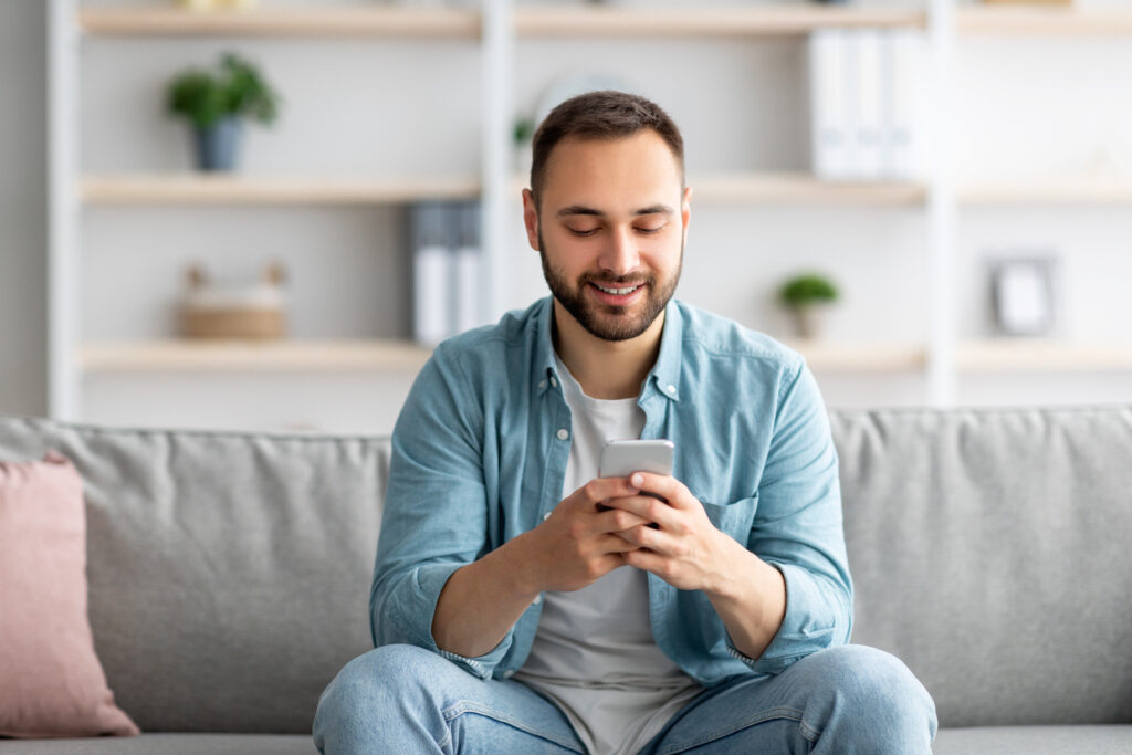 Positive young man sitting on couch with smartphone and texting to his friend, communicating online indoors. Millennial Caucasian guy writing sms, having video call or surfing web at home